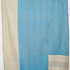 Op de afbeelding: Een blauw en crèmekleurig patchwork quilt. De quilt heeft een groot centraal blauw gedeelte met verticale stiksels en crèmekleurige rechthoekige patches aan de zijkant en onderkant. De randen zijn afgewerkt met rode stiksels.