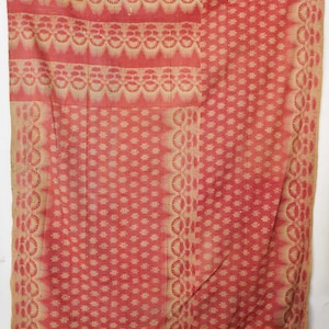 Op de afbeelding: Een rood en beige textiel met een herhalend bloemmotief. De stof heeft rijen gestileerde bloemen en geometrische vormen. Het kleurenpalet is voornamelijk rood en beige, wat een vintage esthetiek creëert. Het textiel lijkt een deken of een decoratieve doek te zijn.