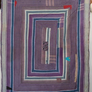 Puede incluir: Un edredón rectangular con un diseño geométrico en tonos morados, azules y blancos. El edredón presenta una serie de rectángulos anidados, con rayas verticales y parches de tela contrastantes. La estética general es vintage y artesanal.