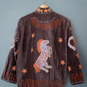 May include: A brown velvet jacket with embroidered details. The jacket features a design of a rearing horse, floral accents, and decorative patterns in orange and gold. The jacket is hanging on a gold hanger.