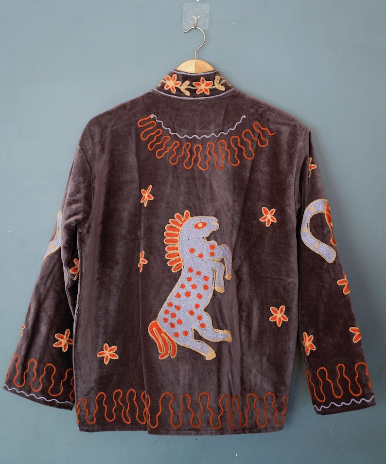 May include: A brown velvet jacket with embroidered details. The jacket features a design of a rearing horse, floral accents, and decorative patterns in orange and gold. The jacket is hanging on a gold hanger.