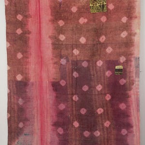 May include: A rectangular textile featuring a deep pink and maroon tie-dye pattern with small white diamond shapes. The fabric displays visible stitching throughout and includes two small, rectangular patches with dark green and yellow floral prints.
