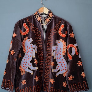 May include: A brown velvet jacket with embroidered horses, horseshoes, and floral designs in orange and lavender. The jacket has a mandarin collar and a black and white striped lining. The jacket is hanging on a wooden hanger.