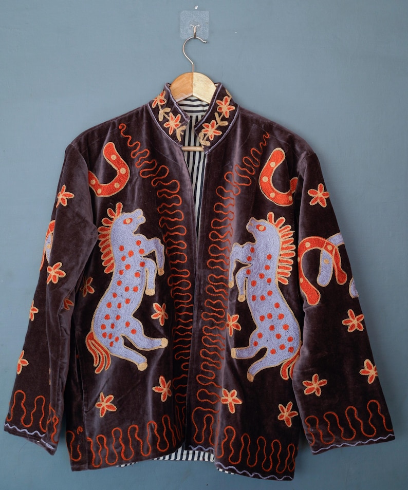 May include: A brown velvet jacket with embroidered horses, horseshoes, and floral designs in orange and lavender. The jacket has a mandarin collar and a black and white striped lining. The jacket is hanging on a wooden hanger.