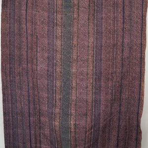 Puede incluir: Textil rectangular con un estampado floral y rayas verticales en tonos morados, marrones y grises. Los bordes están bordeados con un morado más oscuro. El textil es probablemente una manta o un paño decorativo.