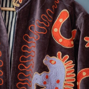 May include: A dark purple velvet jacket with embroidered details. The jacket features orange horseshoe and horse designs, along with orange wavy lines. The collar and cuffs have floral embroidery. The interior lining is striped.