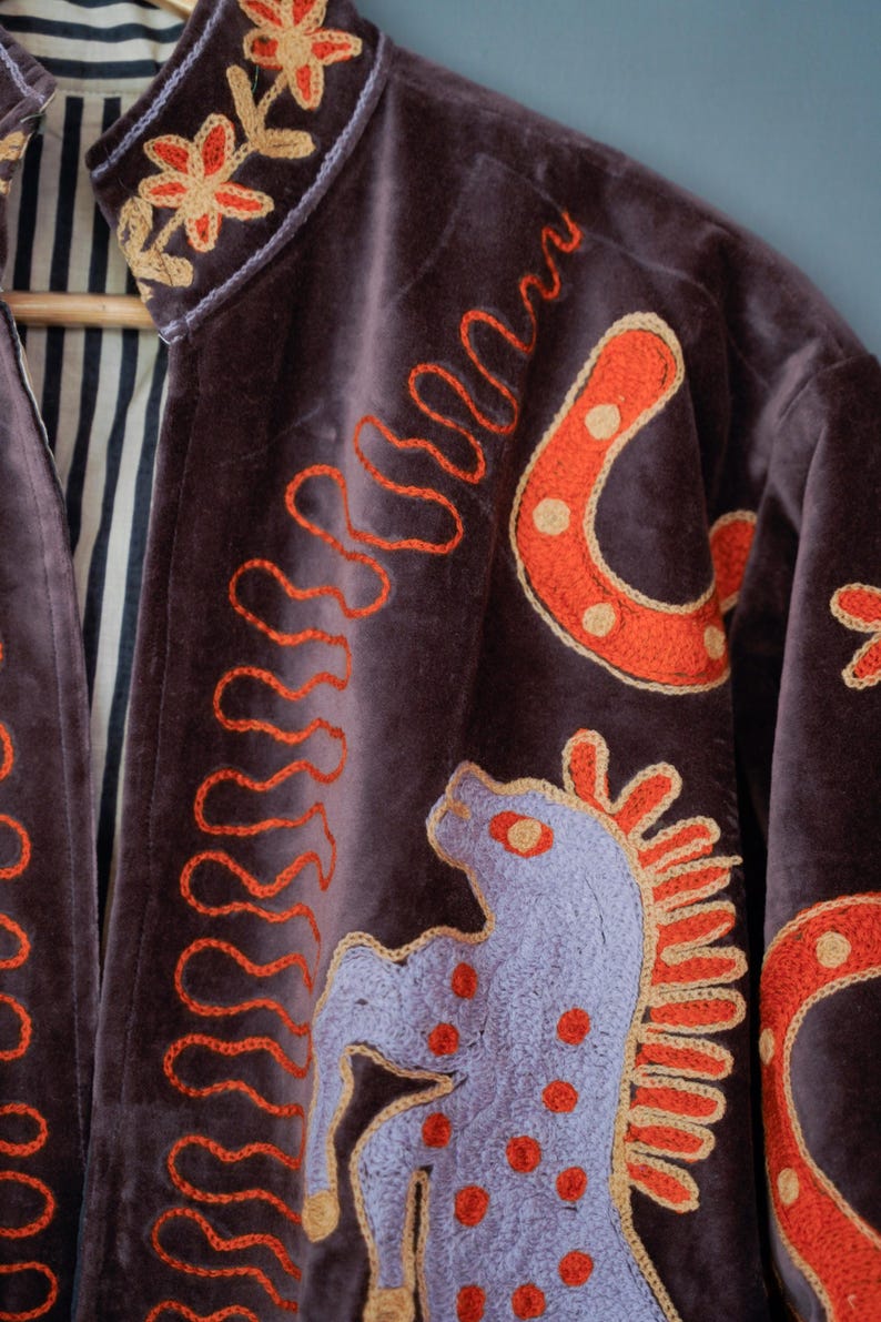 May include: A dark purple velvet jacket with embroidered details. The jacket features orange horseshoe and horse designs, along with orange wavy lines. The collar and cuffs have floral embroidery. The interior lining is striped.