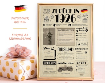May include: A framed print with a gold border, featuring a vintage newspaper design with the text "ZURÜCK IN 1926". The print includes German text, illustrations, and a German flag. A gift box with a pink bow is in the foreground.