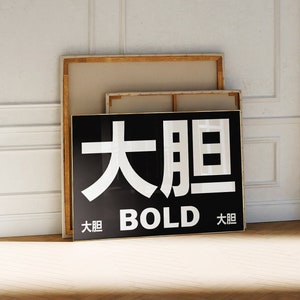 May include: A black and white framed art piece with the word "BOLD" in English and Chinese characters. The artwork leans against a white wall with other framed canvases. The room has a hardwood floor.