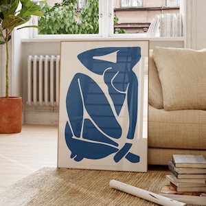 Può includere: Una stampa incorniciata di una figura astratta blu in posizione seduta. La stampa è appesa a una parete bianca in un soggiorno con un divano beige, un tavolino da caffè in legno e una pianta in vaso.