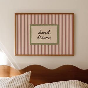 May include: A framed print with the words "Sweet dreams" in a decorative font, set against a striped background. The frame is brown, and the bedding below features striped patterns in neutral tones. The headboard is a dark wood with a wavy design.
