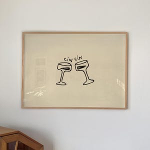 Puede incluir: Una obra de arte enmarcada con dos copas de champán estilizadas brindando. Las palabras "cin cin" están encima de las copas. La obra de arte está en un marco de color claro y cuelga de una pared blanca. Un mueble de madera está en primer plano.