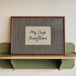 Può includere: Un'opera d'arte incorniciata con una cornice in legno marrone, con uno sfondo a righe e la scritta "My Cup Overflows" in stile manoscritto. L'opera poggia su una mensola verde e color legno, montata su una parete bianca.