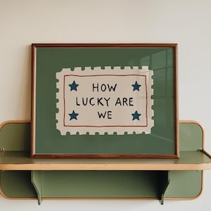 Peut inclure: Une affiche encadrée avec le texte "HOW LUCKY ARE WE" en lettres noires, entourée d'étoiles bleues. L'affiche est posée sur un fond vert et encadrée en marron. Elle repose sur une étagère verte et beige.