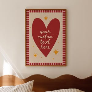 Puede incluir: Un cuadro enmarcado con un gran corazón rojo y el texto "your custom text here" en estilo manuscrito. El corazón está sobre un fondo crema con acentos de estrellas amarillas y un borde a rayas rojas y blancas. El marco es marrón.