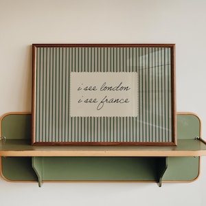 Op de afbeelding: Een ingelijste print met de tekst "i see london i see france" op een gestreepte achtergrond, tentoongesteld op een groen en houten plankje. De plank is aan een witte muur bevestigd, wat een minimalistische esthetiek creëert.