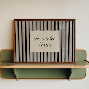 May include: A framed print with the text "love like Jesus" on a striped background. The frame is brown, and the print is displayed on a green and wood-toned shelf. The shelf is mounted on a white wall.