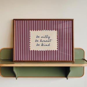 May include: A framed print with a red and white striped background and the words "be silly, be honest, be kind" on a cream-colored, scalloped-edge label. The print rests on a green and wood-toned shelf.