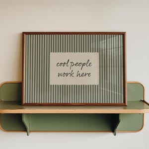 Puede incluir: Un marco con la frase "cool people work here" en una estantería de pared verde y marrón. La impresión tiene un fondo a rayas con una etiqueta blanca. La estantería es de color verde claro.