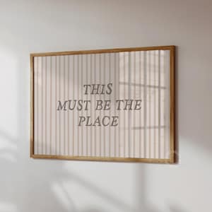 Puede incluir: Un cuadro enmarcado con el texto "THIS MUST BE THE PLACE" en una fuente serif. La obra de arte presenta un fondo a rayas beige y blanco y un marco de madera natural. Está montado en una pared blanca.