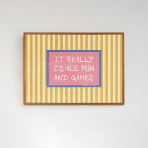 Pode incluir: Uma impressão emoldurada com um fundo listrado amarelo e branco. A impressão apresenta um retângulo rosa com o texto "IT REALLY IS ALL FUN AND GAMES" em branco, emoldurado por uma borda azul. A moldura é marrom.