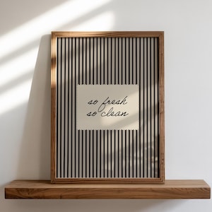May include: A framed print with a wooden frame and a black and white striped background. The print features a cream-colored rectangle with the words "so fresh so clean" in a handwritten font. The print is displayed on a wooden shelf.
