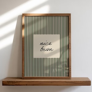 Op de afbeelding: Een ingelijste print met een houten frame en de woorden "nice bum" op een crèmekleurig vierkant. De achtergrond heeft verticale groene en witte strepen. De print wordt weergegeven op een houten plank tegen een witte muur.