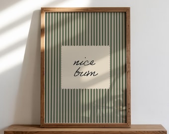 Op de afbeelding: Een ingelijste print met een houten frame en de woorden "nice bum" op een crèmekleurig vierkant. De achtergrond heeft verticale groene en witte strepen. De print wordt weergegeven op een houten plank tegen een witte muur.
