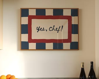 May include: A framed artwork with the text "Yes, Chef!" is displayed on a white wall. The artwork has a red and white center with a blue and white checkered border. A terracotta bowl filled with oranges and two wine bottles are on the counter.