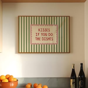 Op de afbeelding: Een ingelijst bord met de tekst "KISSES IF YOU DO THE DISHES" hangt boven een aanrecht. Het bord heeft een groen-witte gestreepte achtergrond en een roze rand. Een terracotta kom met sinaasappels en twee wijnflessen staan op het aanrecht.