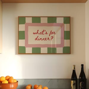 May include: A framed print with the words "what's for dinner?" in red script, set against a pink and green checkered background. A terracotta bowl of oranges and two wine bottles are on a countertop.