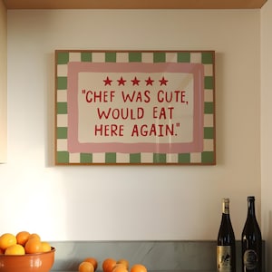 Op de afbeelding: Een ingelijst bord met de tekst "CHEF WAS CUTE, WOULD EAT HERE AGAIN." wordt weergegeven op een witte muur. Het bord heeft een roze en groene rand. Een terracotta kom gevuld met sinaasappels staat op een aanrecht, samen met verschillende losse sinaasappels en twee wijnflessen.