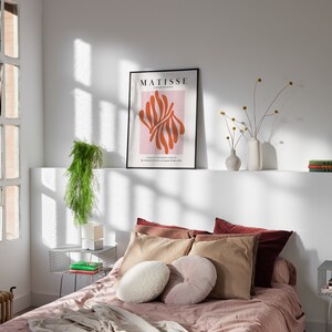 May include: A bedroom with a bed with a pink blanket and brown pillows. There is a framed poster on the wall that says "MATISSE" with an abstract design in orange and pink. There are two side tables with books and a plant on one of them.
