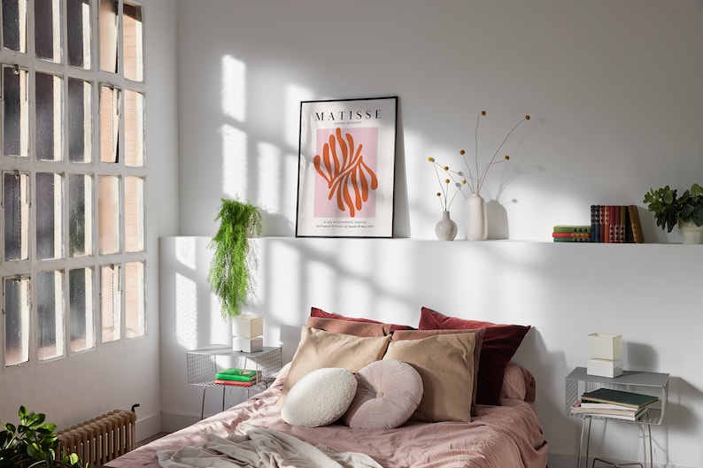 May include: A bedroom with a bed with a pink blanket and brown pillows. There is a framed poster on the wall that says "MATISSE" with an abstract design in orange and pink. There are two side tables with books and a plant on one of them.