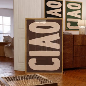 Puede incluir: Grandes impresiones artísticas enmarcadas con la palabra "CIAO" en letras de bloque beige sobre un fondo marrón oscuro. Se ven dos impresiones enmarcadas adicionales con la misma palabra, sobre fondos negro y verde. Las impresiones se exhiben en una habitación con suelo de madera.