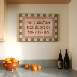 May include: A rectangular wall print in a wooden frame featuring a green and white checkered border, a pink rectangular accent, and the red text: YOUR CUSTOM TEXT, QUOTE OR SONG LYRICS. The print is displayed above a counter with oranges.