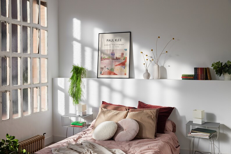 May include: A bedroom with a bed with a pink duvet and a white and brown pillow. There is a framed poster on the wall that says "PAUL KLEE" and a plant on a side table.
