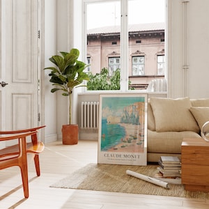 May include: A framed art print of a painting by Claude Monet titled "The Beach and the Palette of Sunset, 1897". The print is hanging on a white wall in a living room with a beige sofa, a wooden chair, and a potted plant.