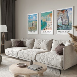 May include: Three framed art prints of seascapes by Claude Monet. The prints are hung on a light beige wall above a white couch. The prints are all in white frames with a white mat.