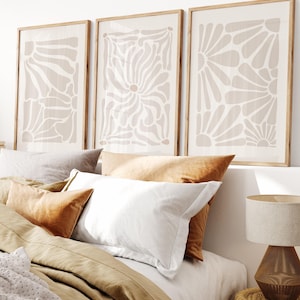 Peut inclure: Un ensemble de trois cadres muraux présentant des motifs floraux abstraits beiges sur fond clair, exposés dans des cadres en bois clair au-dessus d'un lit avec des oreillers superposés blancs, fauves et gris, et une lampe de chevet texturée.