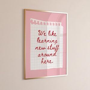 May include: A framed print with a gold-colored frame. The print features a white, grid-lined paper with the text "We like learning new stuff around here" in red cursive lettering, set against a pink background.