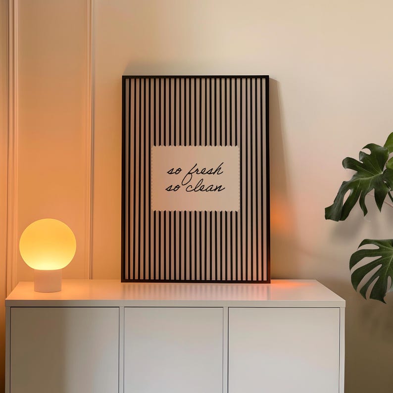 May include: A framed art print with a black and white striped background and the words "so fresh so clean" in a handwritten font. The print is displayed on a white cabinet, with a round, glowing lamp to the left.