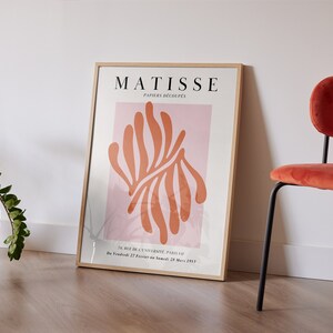 May include: A framed poster with the text "MATISSE PAPIERS DECOUPES" and an abstract orange and pink design. The poster is hanging on a white wall next to a red velvet chair.