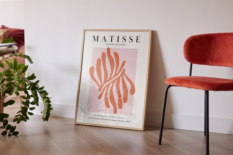 May include: A framed poster with the text "MATISSE PAPIERS DECOUPES" and an abstract orange and pink design. The poster is hanging on a white wall next to a red velvet chair.