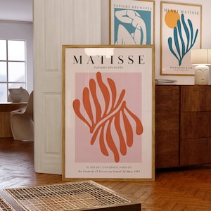May include: A pink and orange poster with the text "MATISSE PAPIERS D&Eacute;COUP&Eacute;S" and a large orange abstract design. The poster also includes the text "70, RUE DE L'UNIVERSIT&Eacute;, PARIS VII Du Vendredi 27 Fevrier an Samedi 28 Mars 1953".