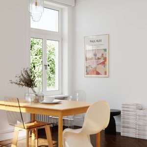 May include: A wooden dining table with four clear plastic chairs. A white chair is pulled out from the table. A stack of magazines is on the floor next to a black side table. A framed print of a painting by Paul Klee hangs on the wall. The painting is titled "Castle and Sun" and is dated 1928.