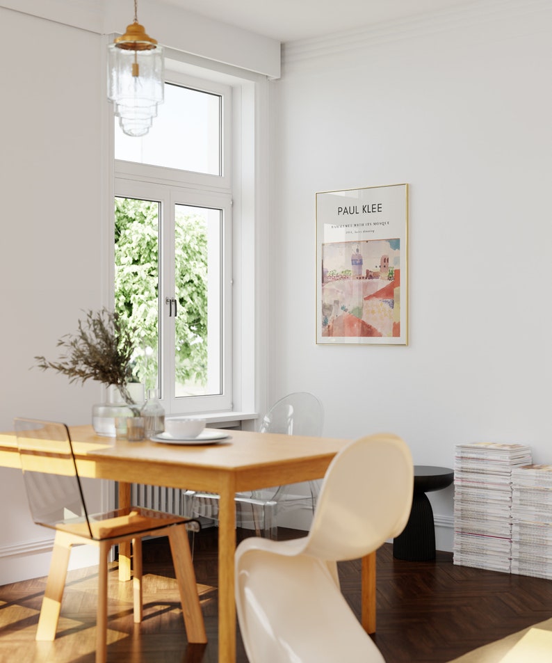 May include: A wooden dining table with four clear plastic chairs. A white chair is pulled out from the table. A stack of magazines is on the floor next to a black side table. A framed print of a painting by Paul Klee hangs on the wall. The painting is titled "Castle and Sun" and is dated 1928.