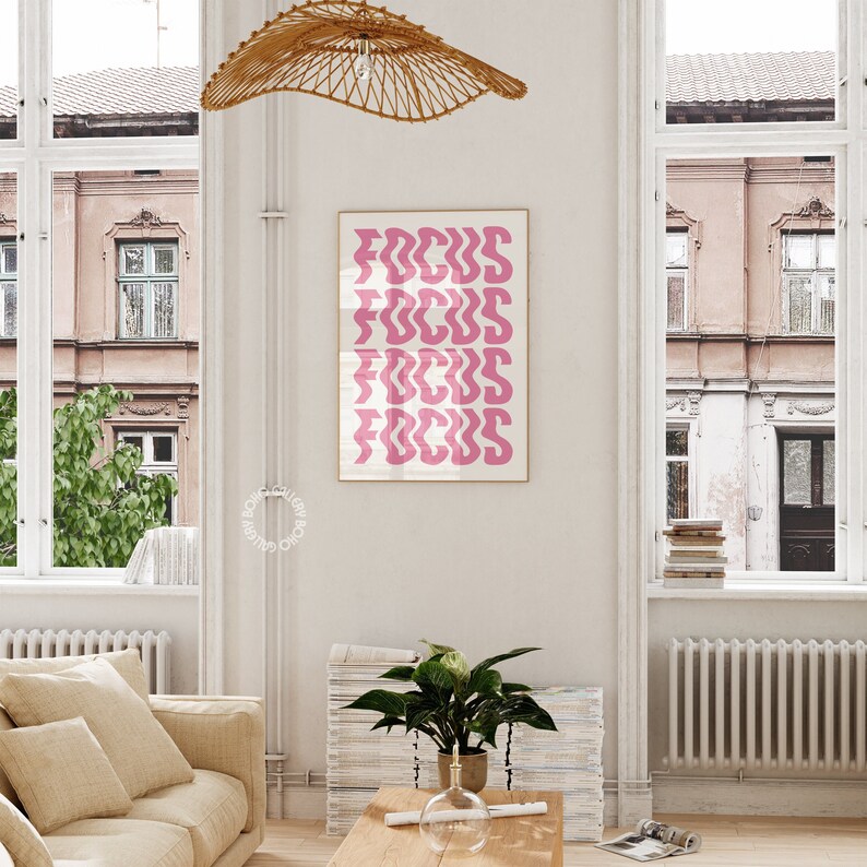 May include: Pink and white print with the word "FOCUS" repeated in a wavy font. The print is framed and hanging on a white wall in a living room setting.