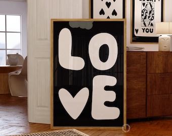 May include: Black and white framed print with the word "LOVE" in large, stylized letters. The letters are white on a black background. The print is hanging on a wall with two other framed prints, one with a playing card and the other with a queen of hearts.