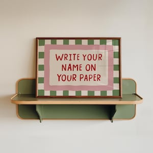 May include: A rectangular framed print featuring a green and white checkered border with a pink inner frame and the red text WRITE YOUR NAME ON YOUR PAPER, resting on a green metal wall shelf with rounded edges and a light wood trim.